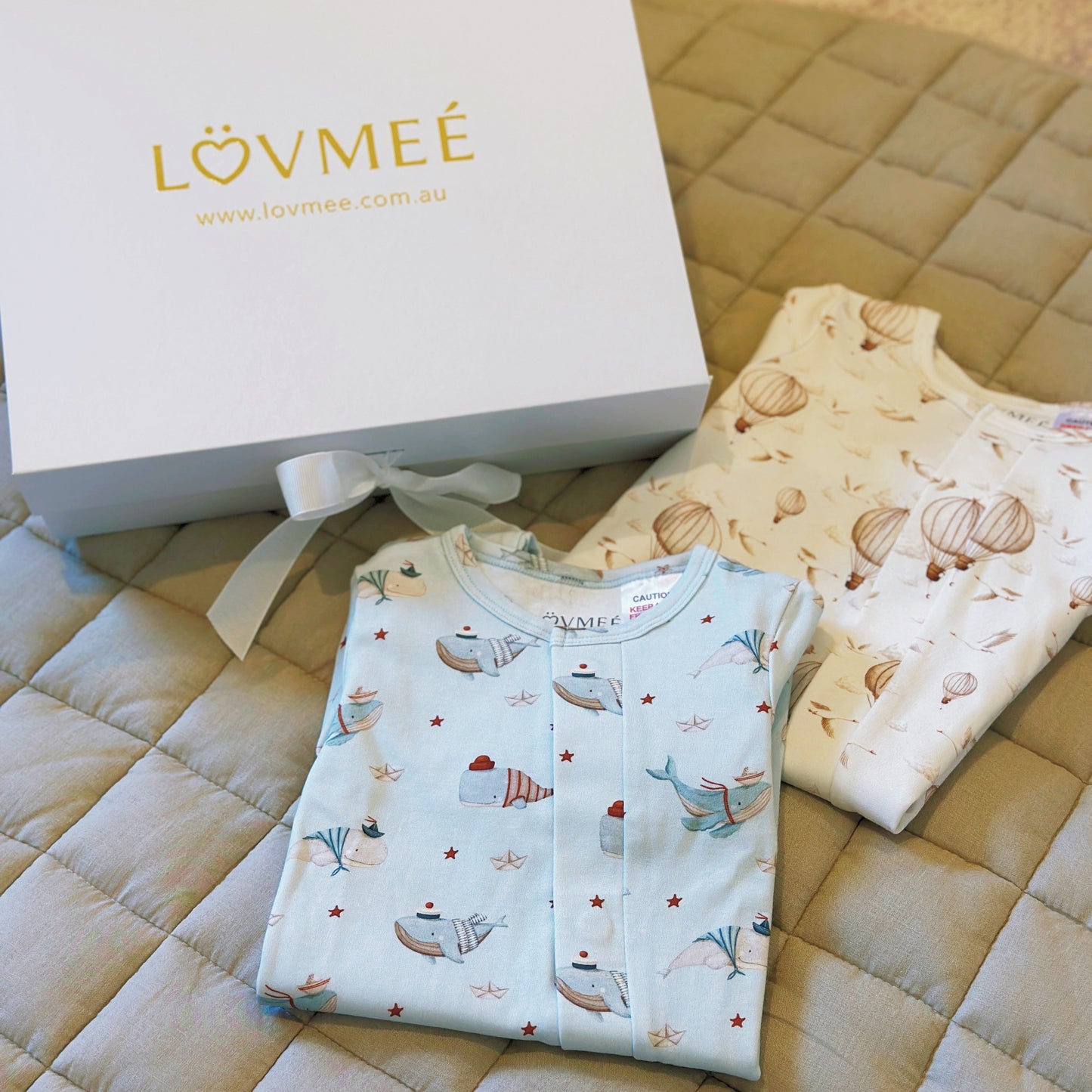 Folded baby clothes with a white box labeled 'LOVME®' on a quilted surface.